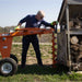 Man operating a Brave hydraulic Towable Auger in an outdoor setting with stacked logs nearby.