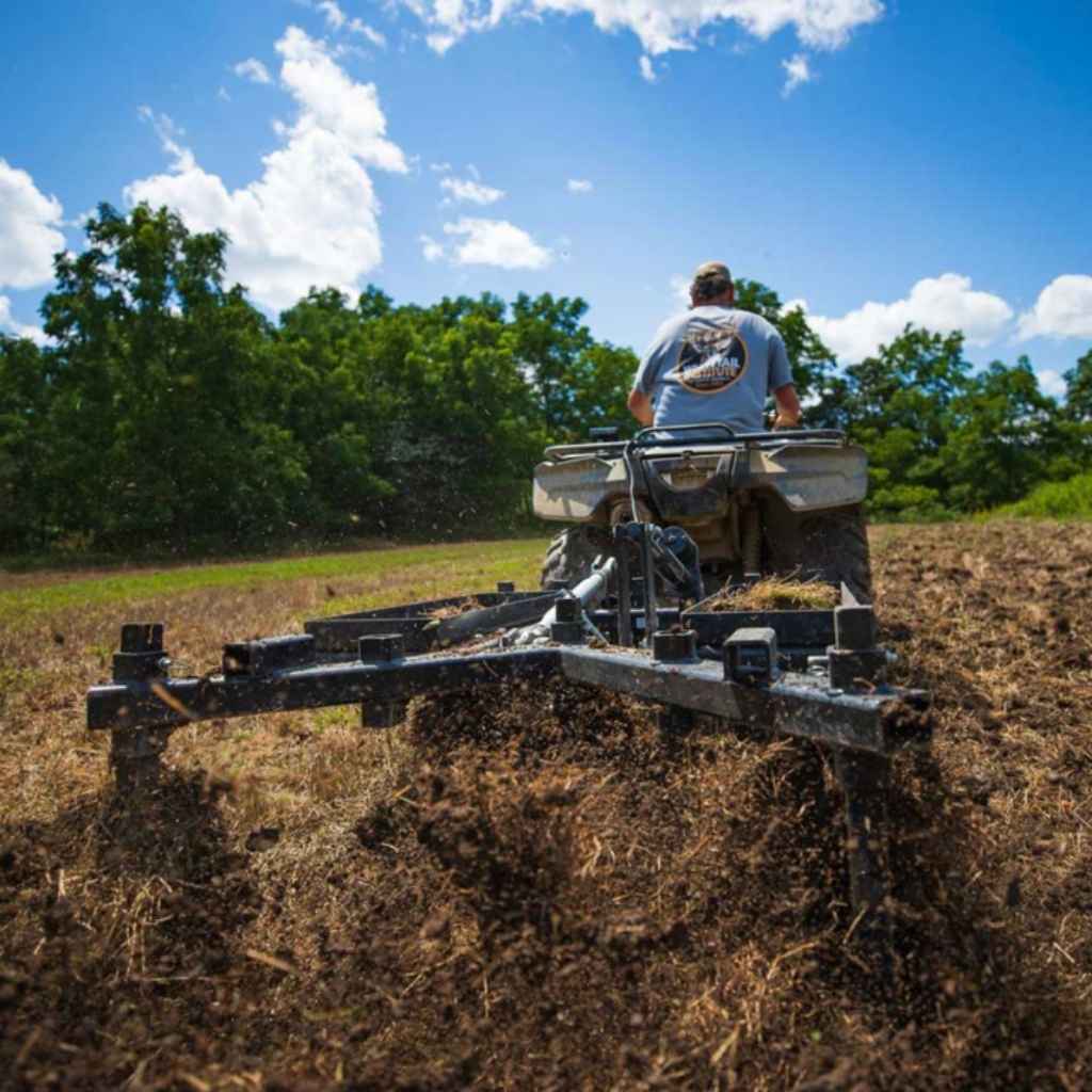 Person operating an ATV with a GreyWolf till n Claw chisel plow & Cultivator attached in a field with trees and blue sky in the background