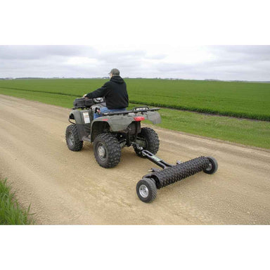 Person riding an ATV with a GreyWolf Cultipacker on a dirt road in a rural setting.