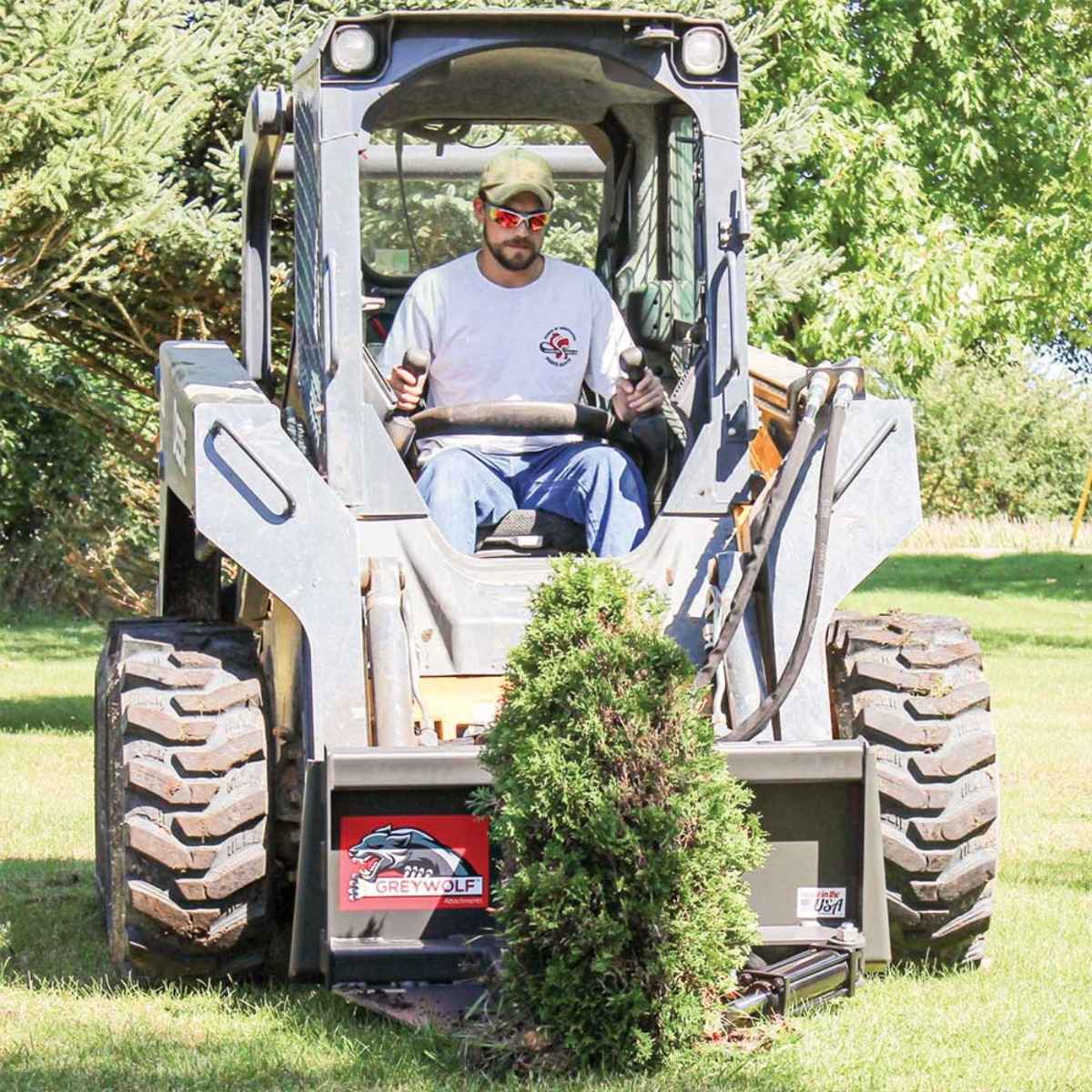 Person operating a skid steer loader with a Greywolf tree puler for skid steer handling a tree on a grassy area