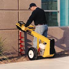Person using a yellow GroundHog 1M5C one-man earth drill on a sidewalk next to a building.