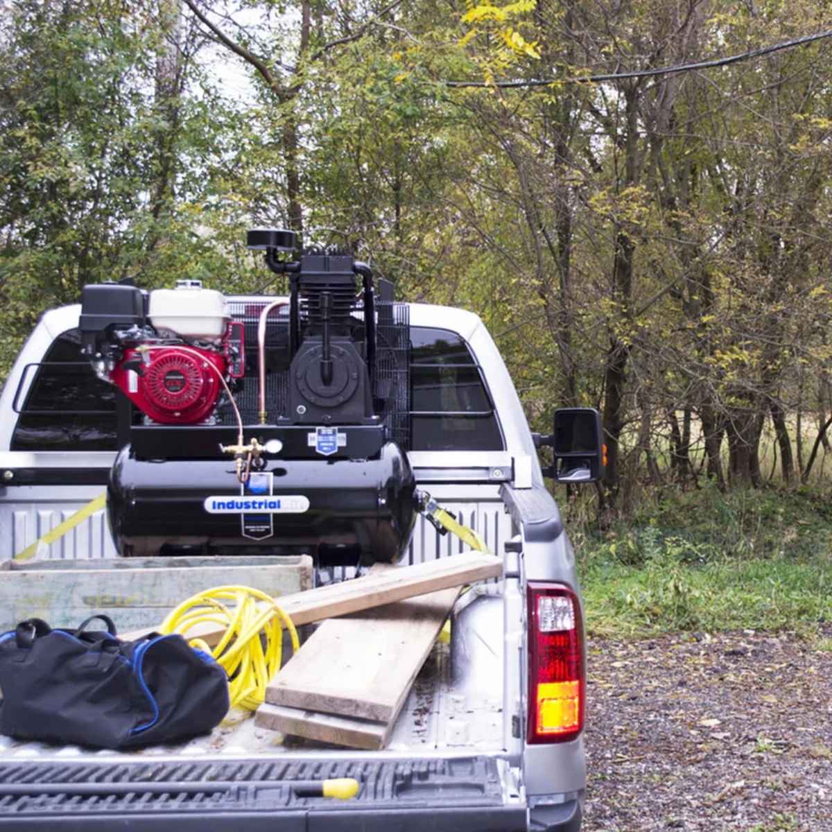 Back of a white truck with a large industrial air 30 gallon belt driven truck mount air compressor and tools, surrounded by trees.