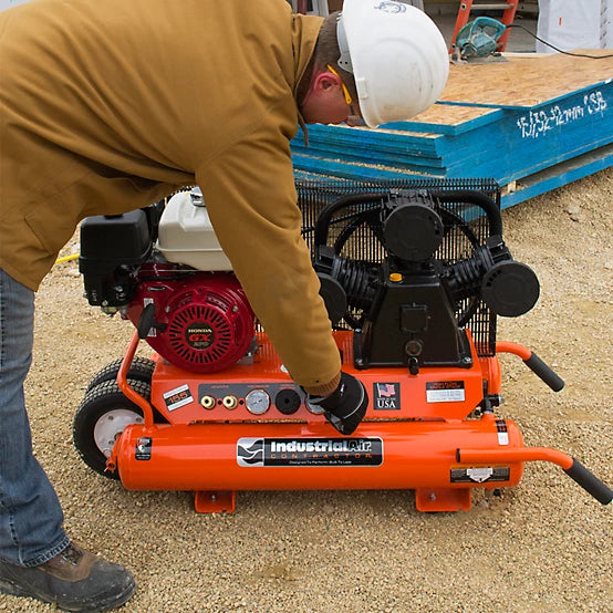 Person working on an orange Industrial Air 9 Gal Gas air compressor with a hard hat on a construction site.