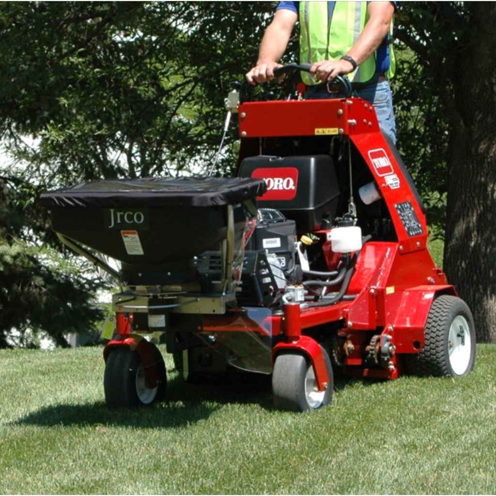 Person operating a jrco 5054 hitch mount spreader in a grassy area with trees in the background