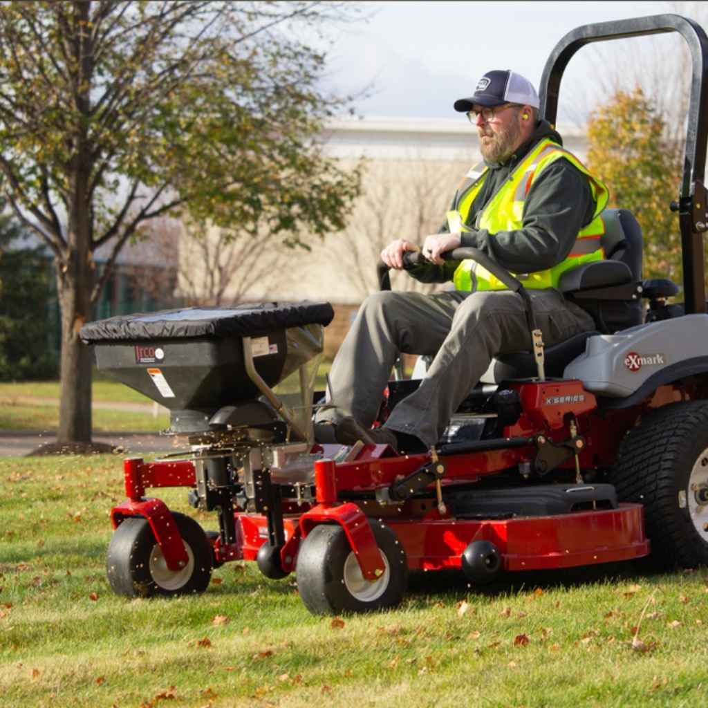 Man operating a lawn mower with a jrco 503 foot control broadcast spreader in an outdoor setting.