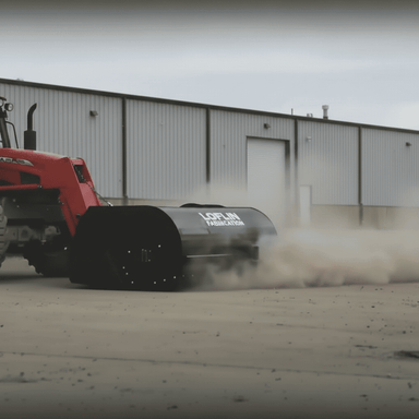 Red tractor with a large black loflin skid steer hydraulic pickup broom labeled 'Loflin' on a dirt surface in front of a building.