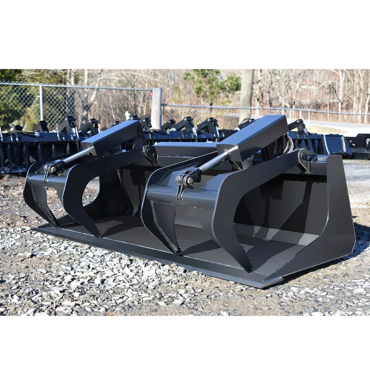 Large Loflin fabrication compact tractor grapple bucket on a gravel surface with a chain-link fence and trees in the background.