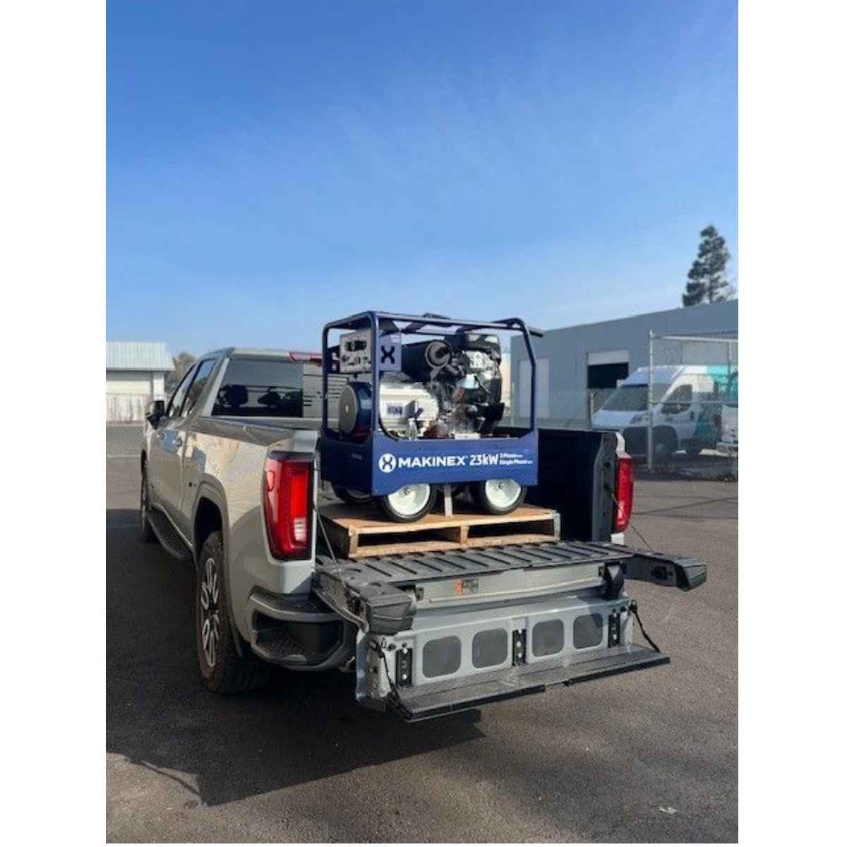 A large silver truck with a Makinex 23 Kw 250v portable generator mounted on a wooden pallet in the bed of a silver pickup truck, parked outdoors on a paved lot.