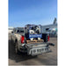A large silver truck with a Makinex 23 Kw 250v portable generator mounted on a wooden pallet in the bed of a silver pickup truck, parked outdoors on a paved lot.