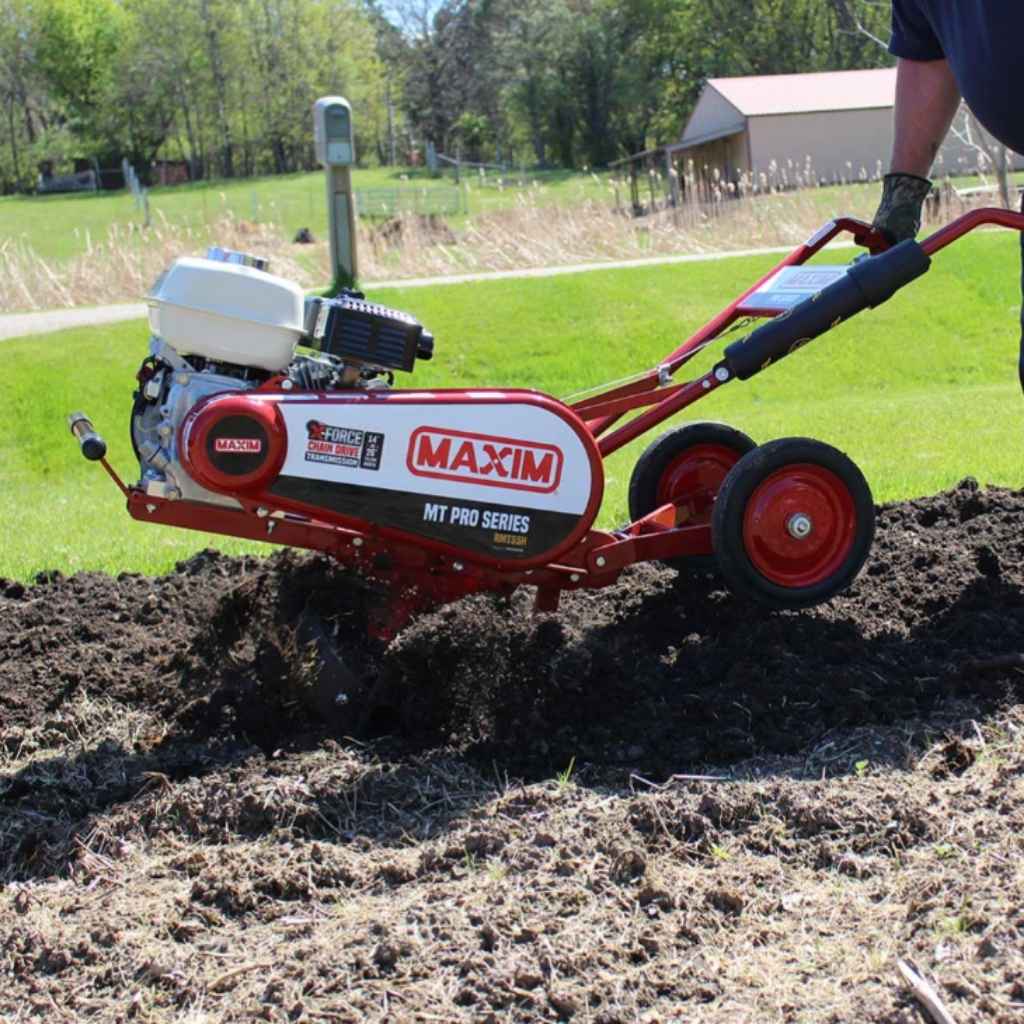 Maxim 26 Inch RMT55H tiller being used on a garden bed with trees and a building in the background