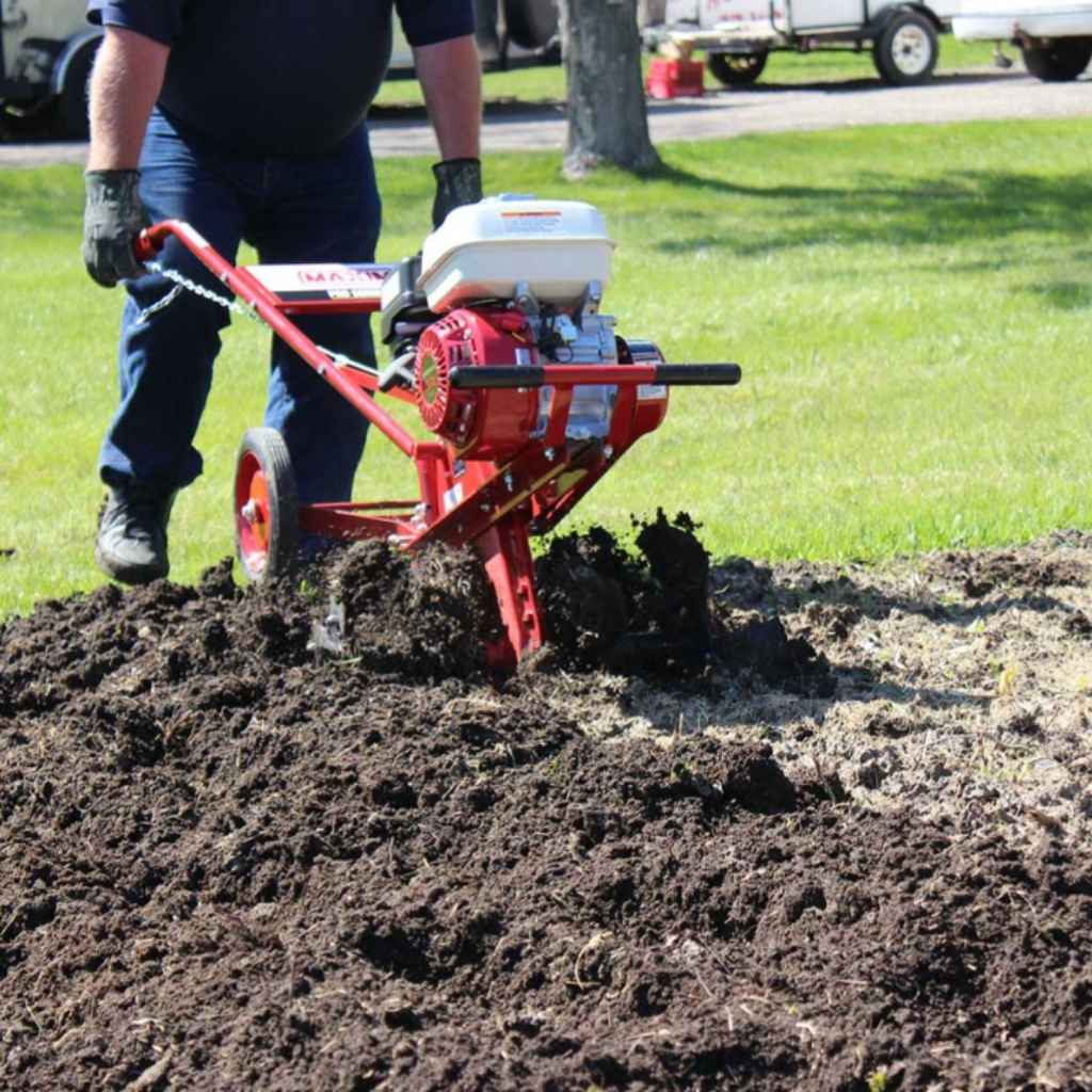 Person using a Maxim 26 inch tiller on a garden bed with grass in the background