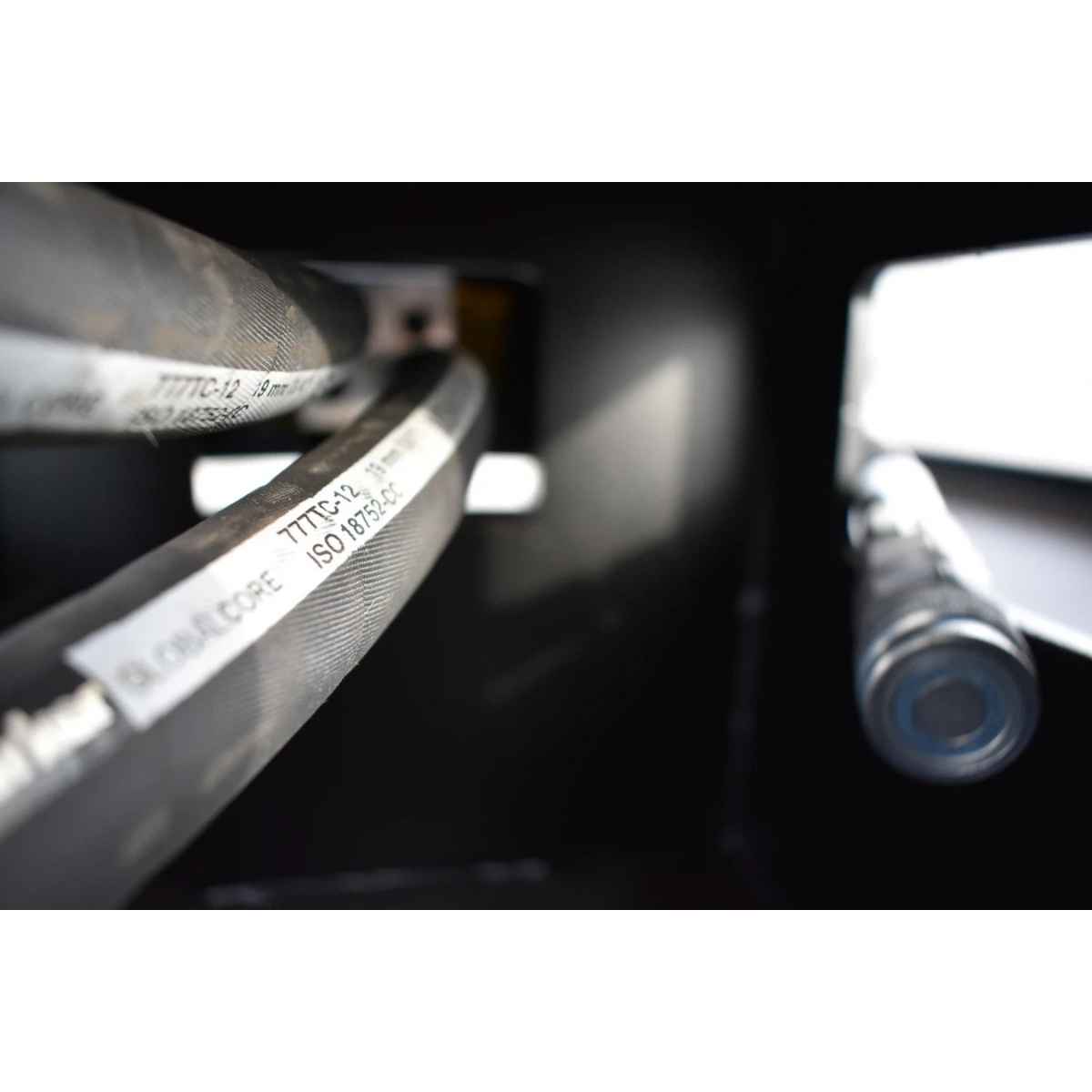 Close-up of black industrial hoses with white printed labels, shown inside a loflin mini excavator brush cutter with a cylindrical metal component on the right.