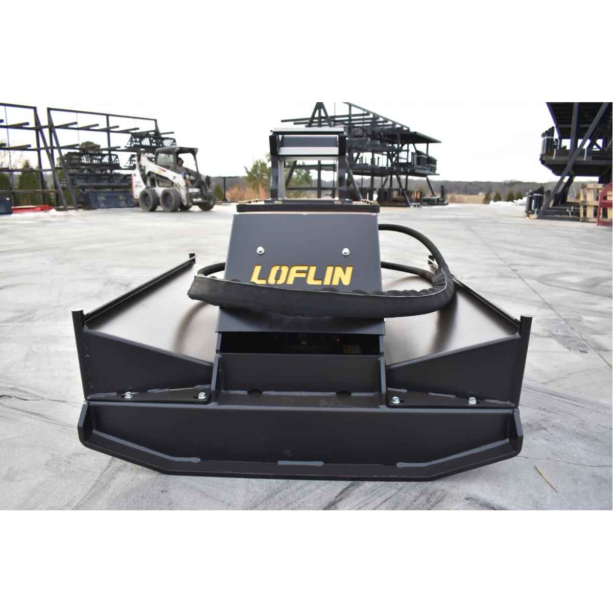 A black mini excavator brush cutter attachment by loflin fabrication, photographed from a front-facing perspective on an outdoor concrete yard with construction equipment in the background.
