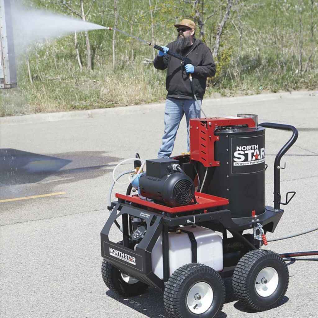 person using a northstar hot water 2750 psi pressure washer on a semi trailer with a concrete ground and trees in the background during the daytime.