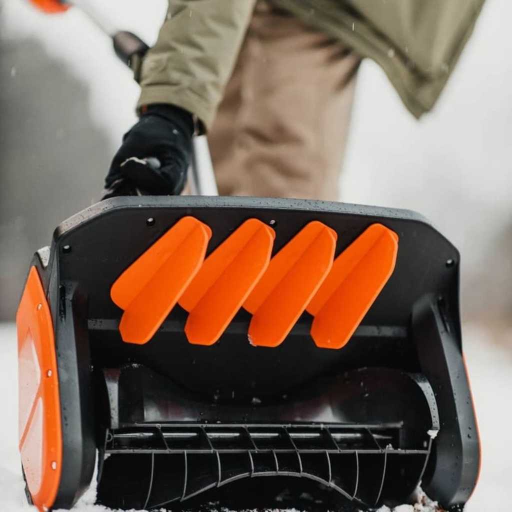 Close-up of a superhandy snow thrower 48v with orange and black design, person operating it in the background.
