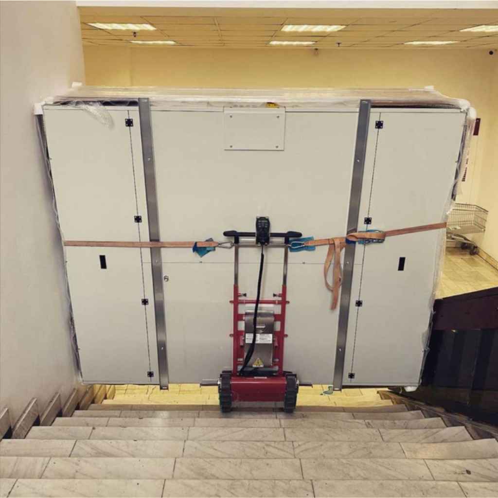 Large white panel with metal straps being transported up a staircase by a zonzini plasmoid stabilized stair climber in an indoor setting