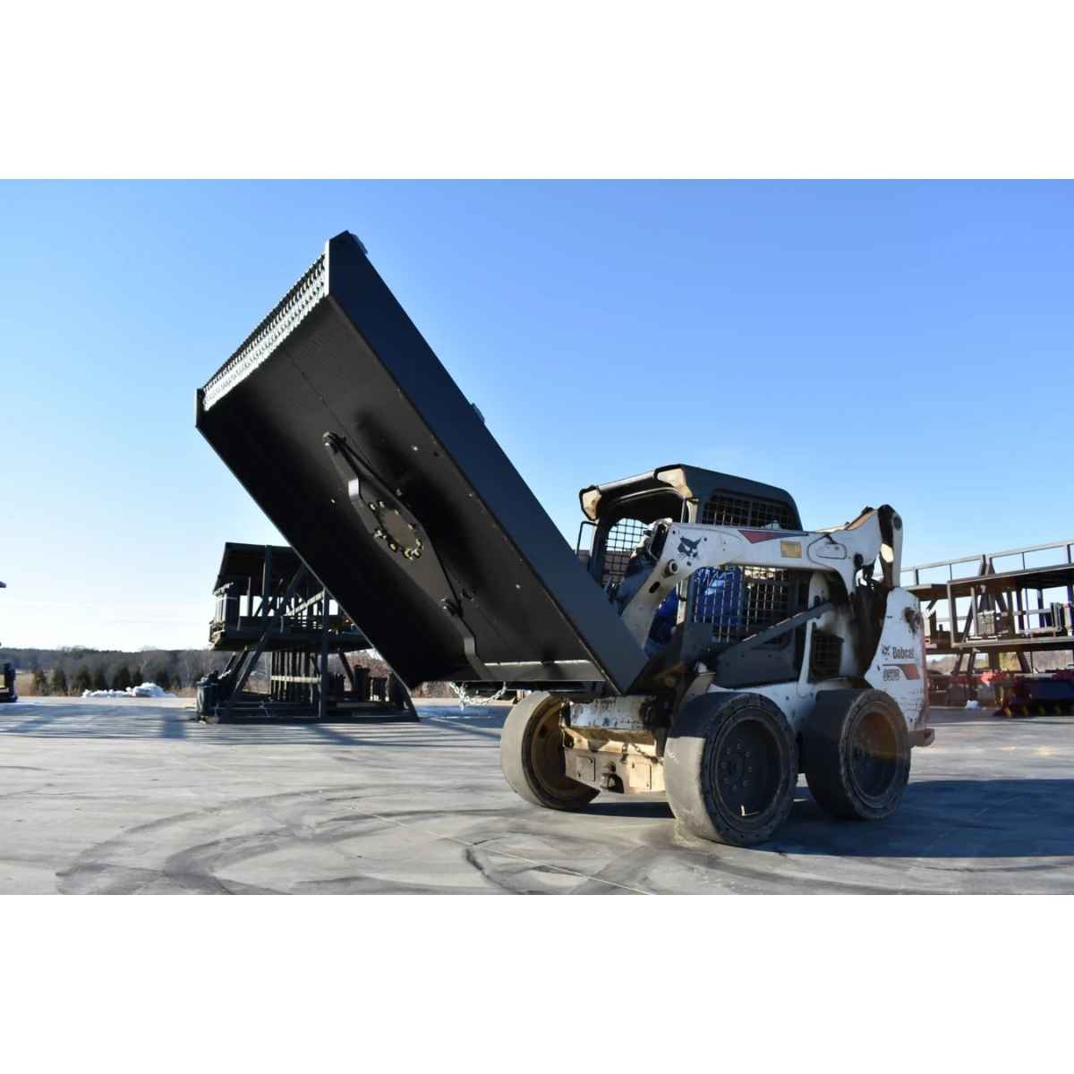A white skid steer loader with a loflin closed front brush cutter attachment is shown outdoors on a paved lot, with the dump bed tilted upward. Other heavy machinery and industrial structures are visible in the background under a clear blue sky.