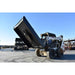A white skid steer loader with a loflin closed front brush cutter attachment is shown outdoors on a paved lot, with the dump bed tilted upward. Other heavy machinery and industrial structures are visible in the background under a clear blue sky.