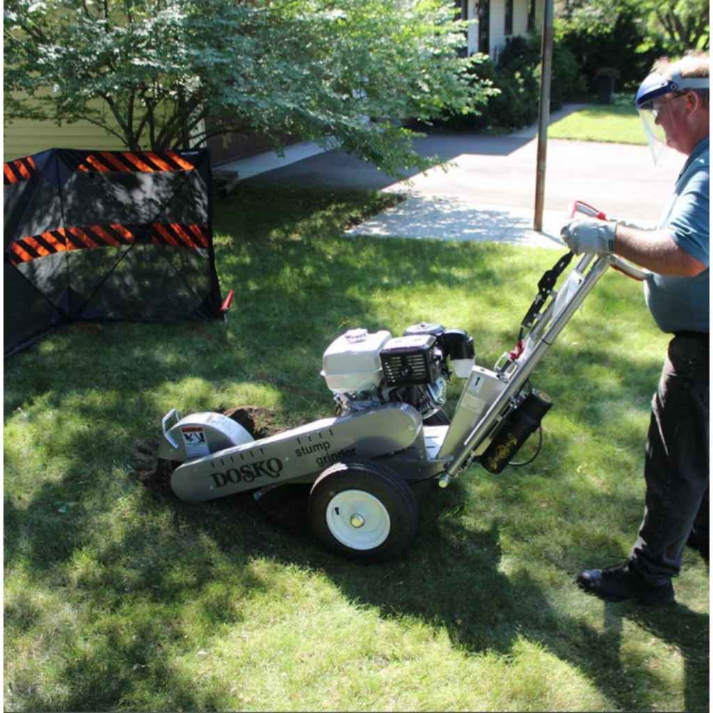Person using a dosko walk behind 13 hp stump grinder on a grassy area with trees and a house in the background
