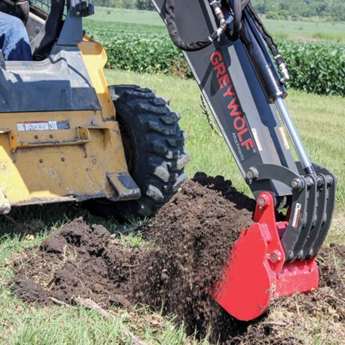 Skid Steer with a GREYWOLF backhoe attachment digging into the ground in an agricultural setting.
