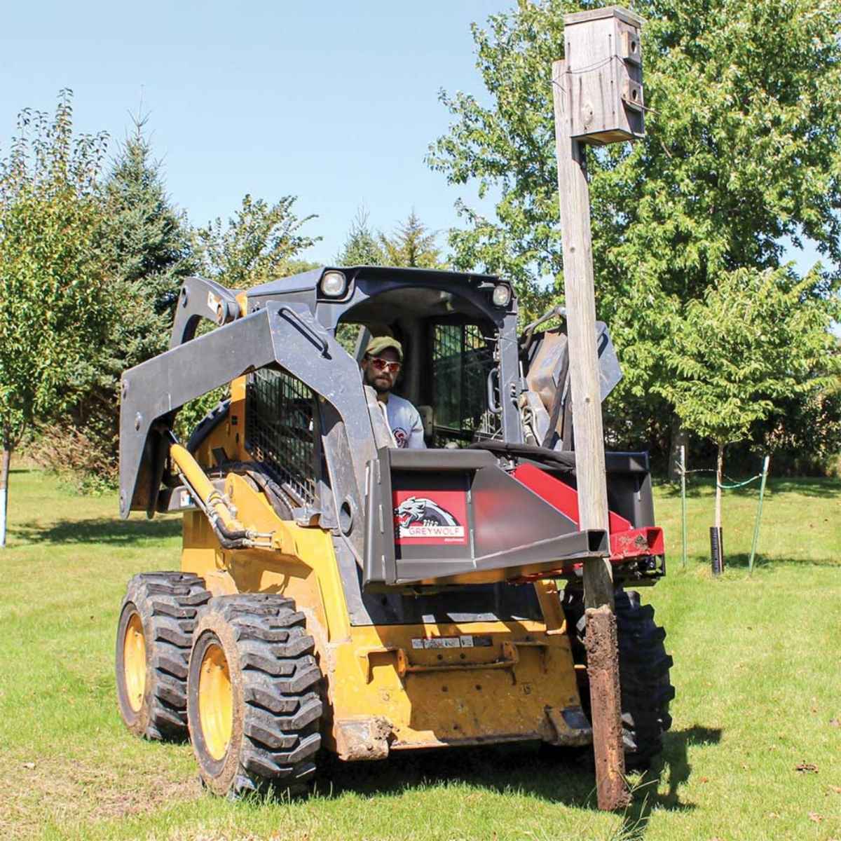 Person operating a skid steer loader with a Greywolf tree puller attachment handling a post in a grassy area.