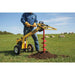 Person using a yellow and red groundhog 1m5c one man auger drill in a grassy field with sheep in the background.