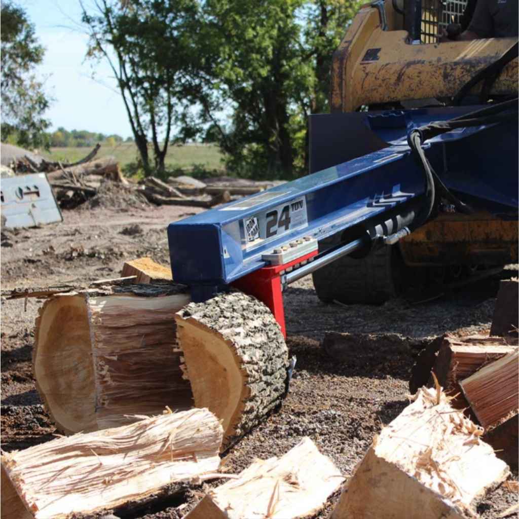 Blue iron and oak 24 ton skid mount log splitter attached to a skid loader splitting a log with trees in the background during the daytime.