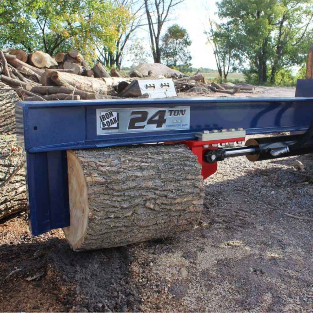 a blue and red iron and oak skid mount 24 ton log splitter holding a log in a wooded area durinf the daytime with logs and trees in the background.