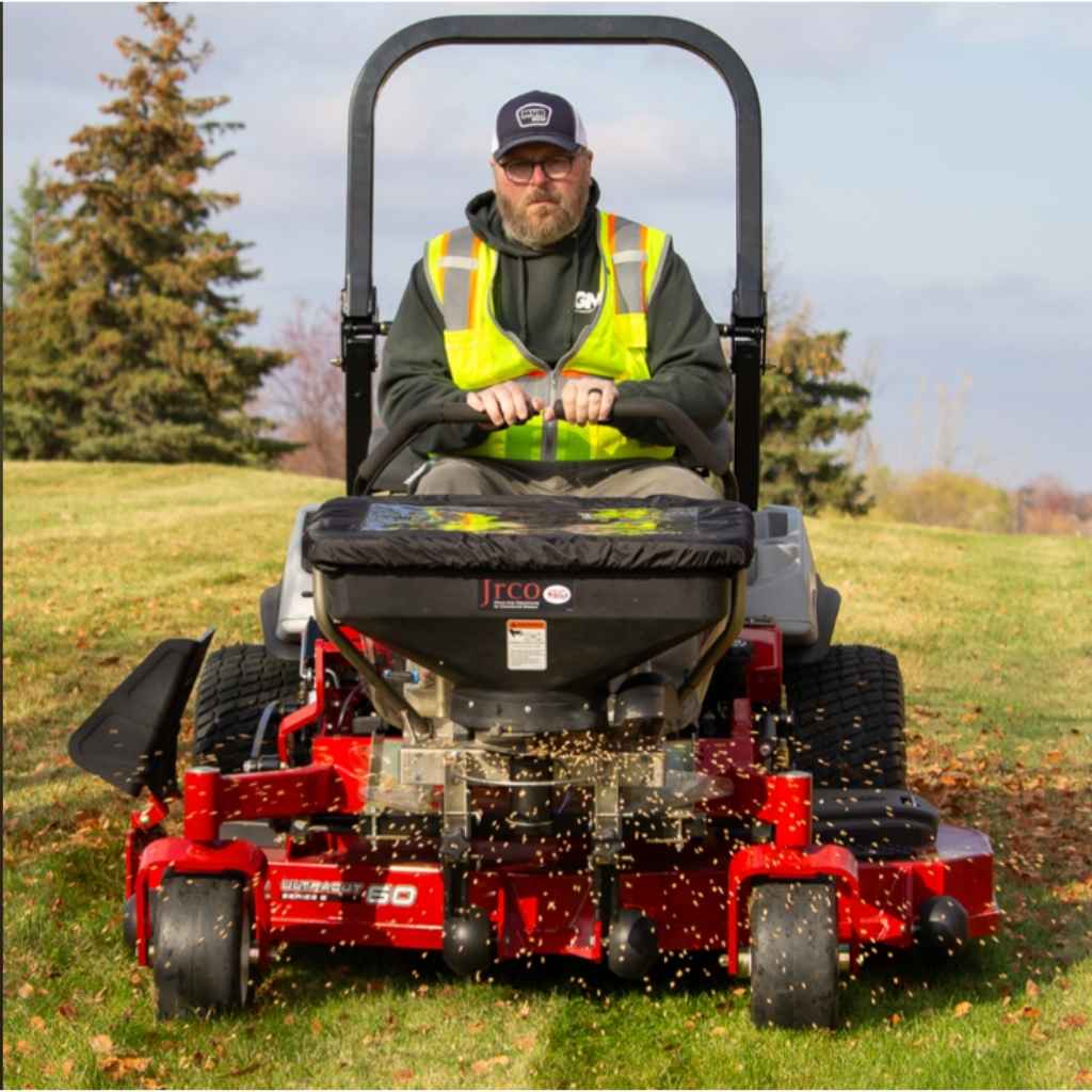Person operating a red lawn mower with a jrco foot control broadcast spreader in an outdoor setting with trees and grass.