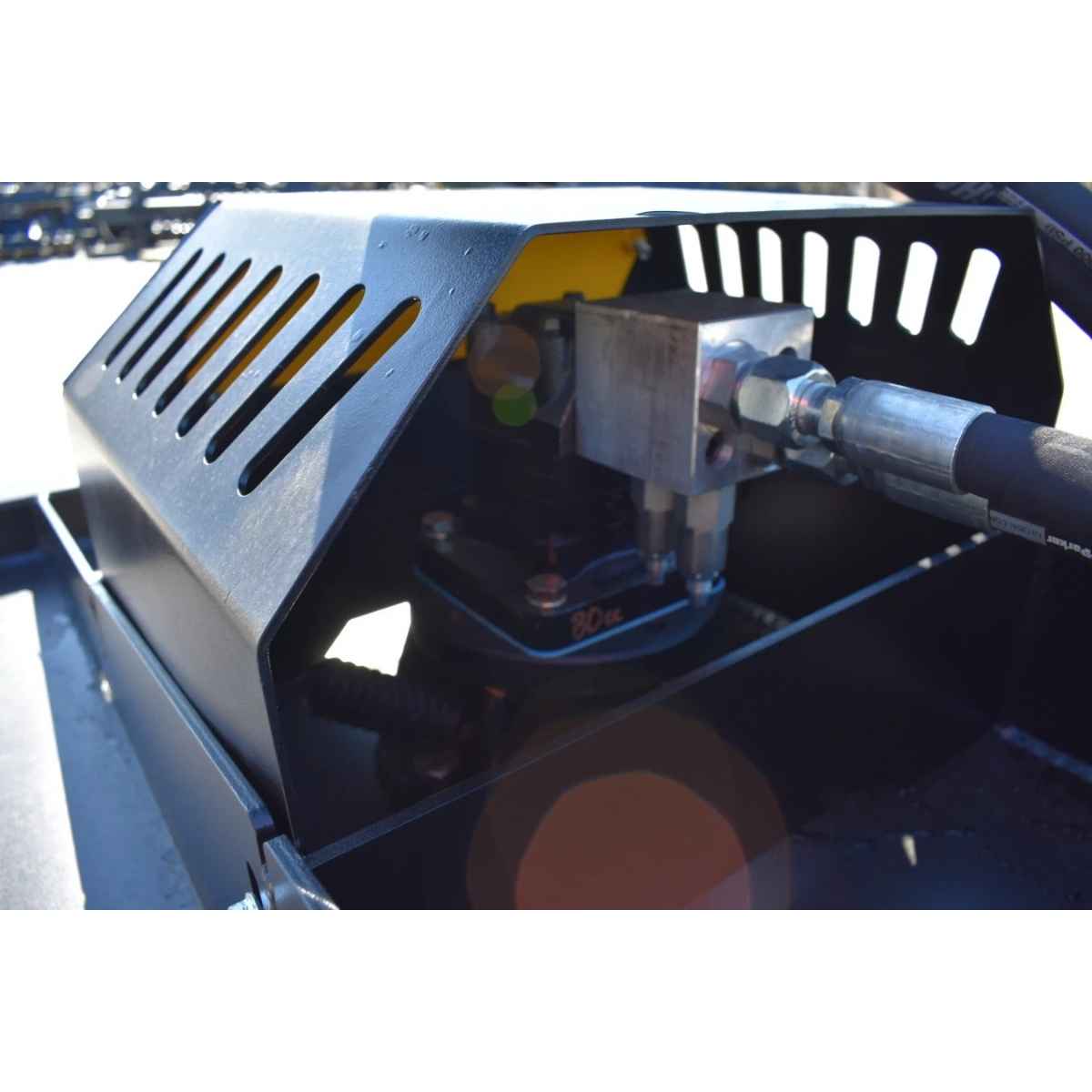 Close-up of a black metal hydraulic power unit with a slotted protective cover to a loflin fabrication brush cutter closed front, showing internal components, hoses, and a metal fitting under bright outdoor light.