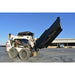 A white skid steer loader with a large black loflin closed front brush cutter attachment raised overhead, parked on a concrete lot with a warehouse building and blue sky in the background.