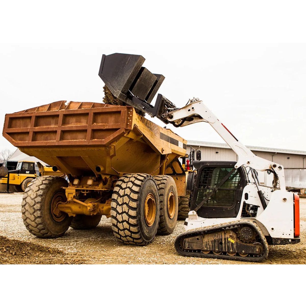 mini skid steer dumping material into dump truck using the loflin front dump bucket on a gravel floor with a factory in the background