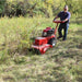 Person operating a red maxim brush cutter 26 inch in a grassy area with trees in the background