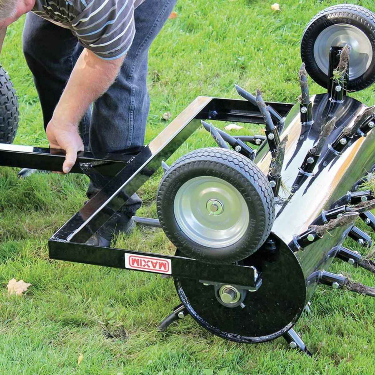 A person holding a black maxim 36 inch tow behind plug aerator with wheels on a grassy field