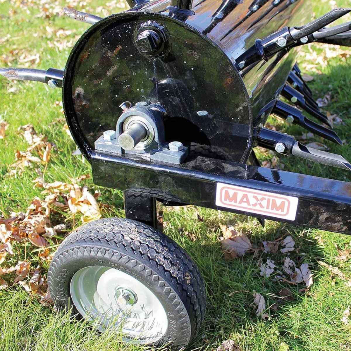 Close-up of a black Maxim 36 inch aerator tow behind with the 'MAXIM' branding on the side of aerator that is on grass with leaves.