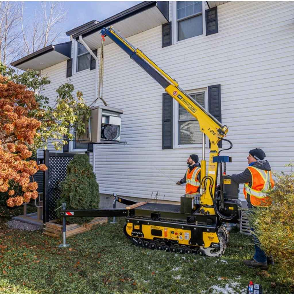 Two workers using a yellow movex mini tracked crawler crane to lift an air conditioner outside a house.