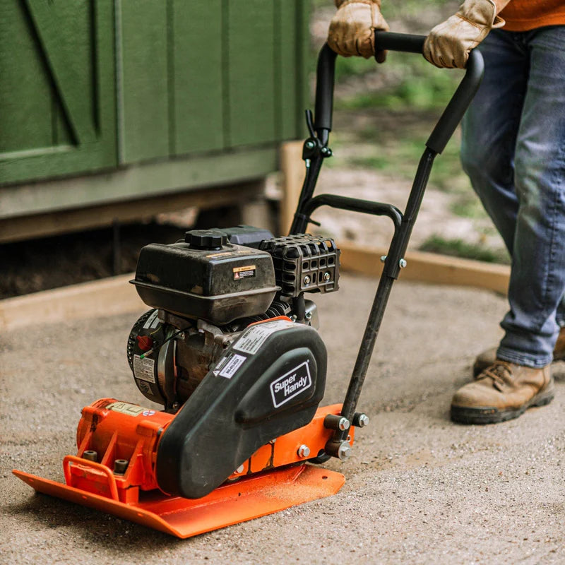 An orange and black SuperHandy vibratory plate compactor with a gas engine, designed for compacting soil and gravel. Engine view outdoors in use.