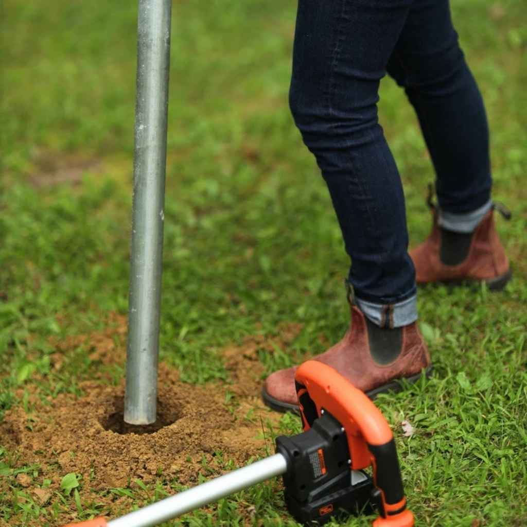 Person using a superhandy electric earth auger 20v to create a hole in the ground with a metal pole in the background.
