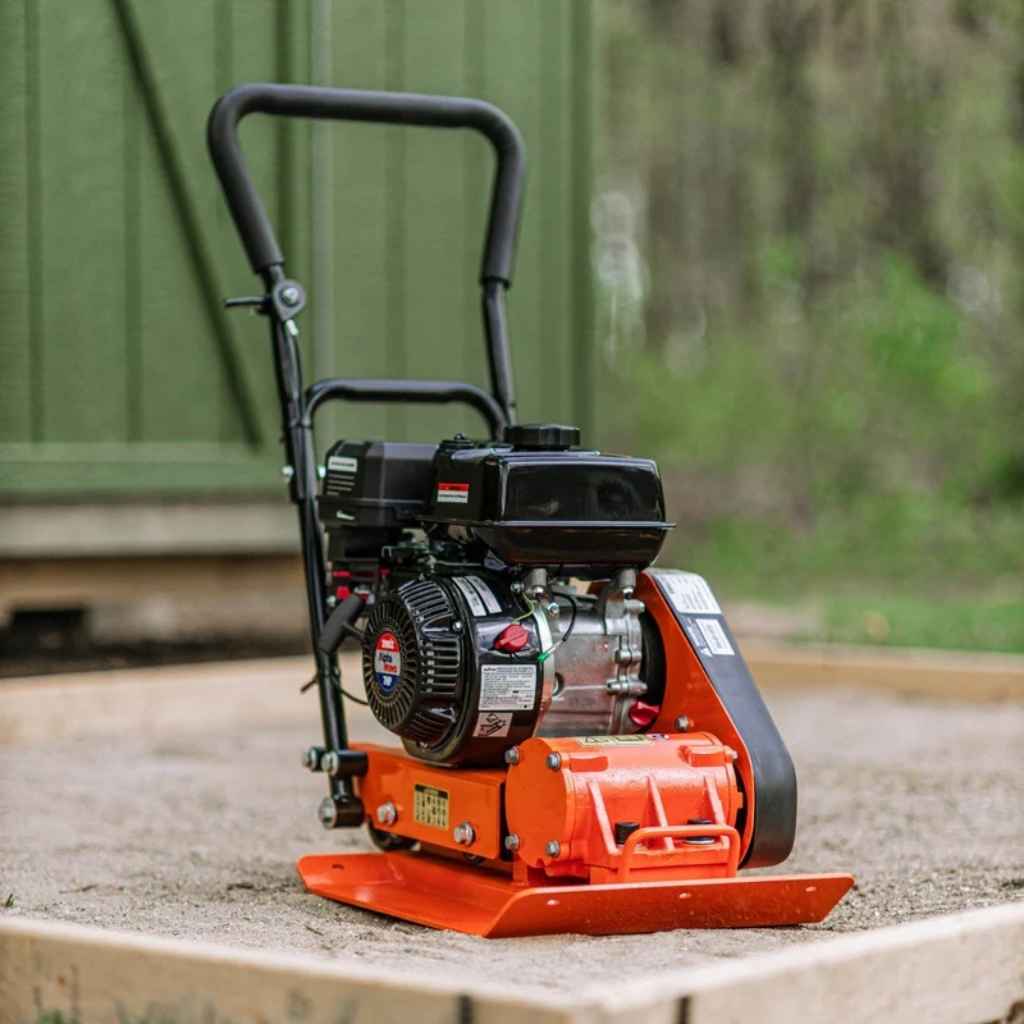 Small orange and black superhandy vibratory plate compactor on a concrete surface with a blurred green background