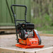 Small orange and black superhandy vibratory plate compactor on a concrete surface with a blurred green background