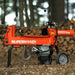 An orange and black 14 Ton Electric Log Splitter with a 20 inch max log length. The equipment is corded and has wheels for mobility. Side view outdoors.