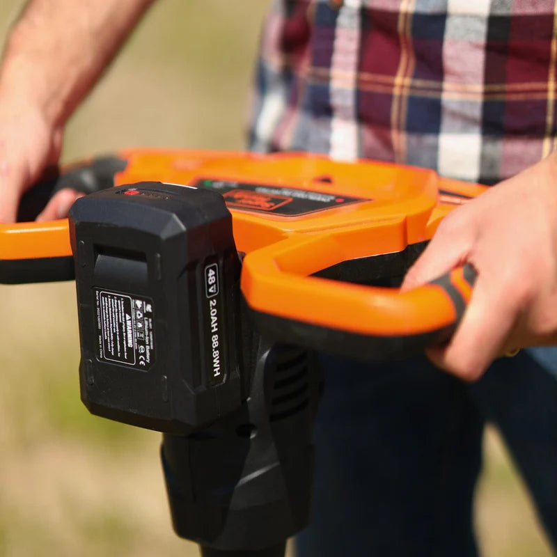 An electric earth auger with a 48V 2Ah battery, featuring an orange and black color scheme and a spiral digging bit. Battery view outoors.
