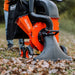 An orange and black gas powered leaf vacuum and wood chipper with a black collection bag and wheels for mobility. Front view outdoors in use with logo showing.