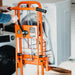 An orange material lift stacker and pallet dolly with a hand crank and swivel caster wheels. Handle view indoors in use.