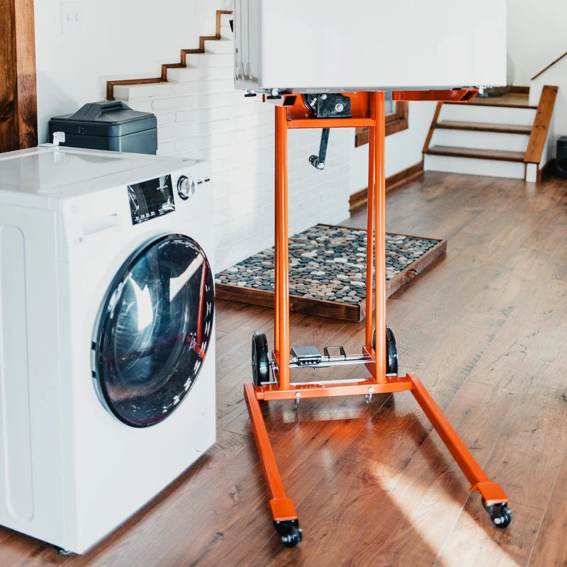 An orange material lift stacker and pallet dolly with a hand crank and swivel caster wheels. Front view indoors.