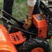 A superhandy 16" rear tine tiller with orange, black, and gray coloring, featuring a gas engine and large wheels for garden soil preparation. Top view outdoors.