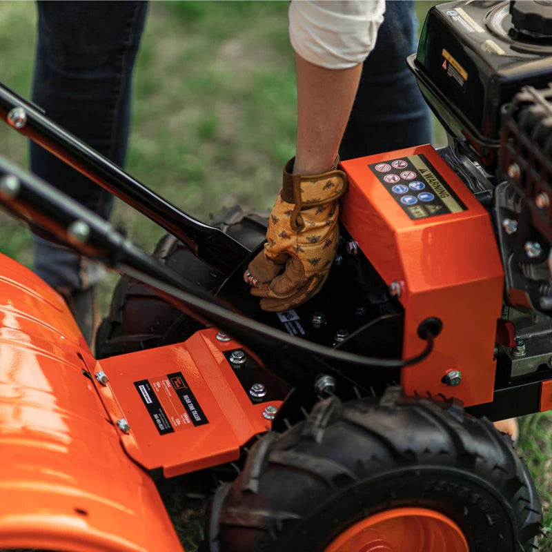 A superhandy 16" rear tine tiller with orange, black, and gray coloring, featuring a gas engine and large wheels for garden soil preparation. Top view outdoors.