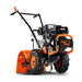 A rear tine tiller with orange, black, and gray coloring, featuring a gas engine and large wheels for garden soil preparation.