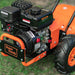A rear tine tiller with orange, black, and gray coloring, featuring a gas engine and large wheels for garden soil preparation. Engine view outdoors.