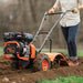 Person using an orange and black Superhandy rear tine tiller 16 inch to till loose soil 
